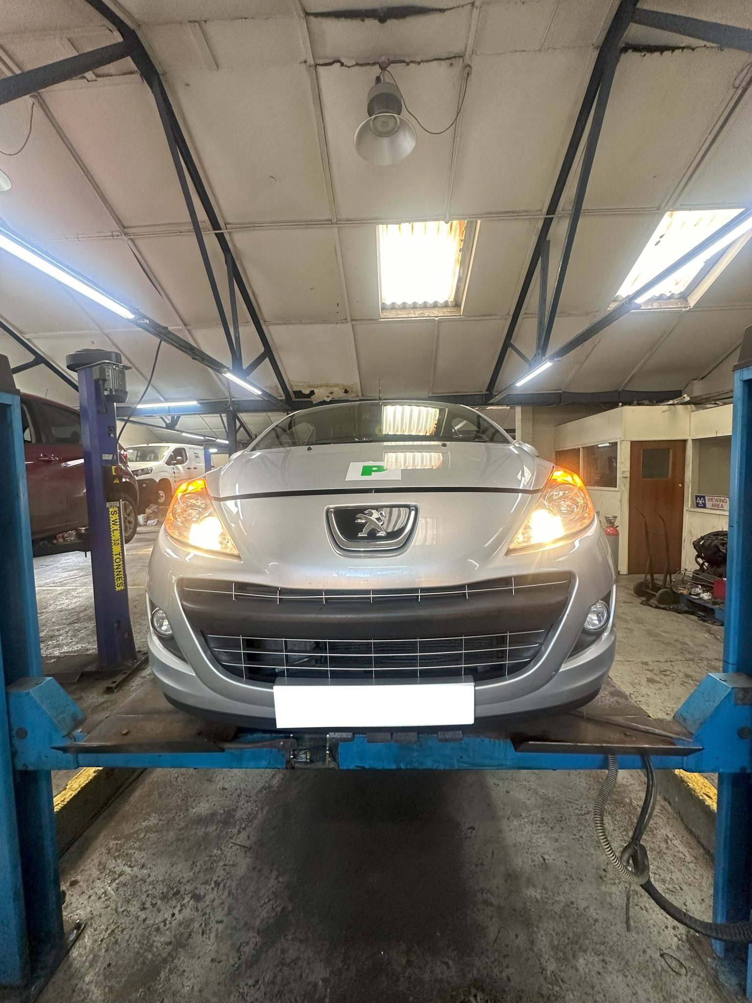 Silver Peugeot car on a lift inside Malling Garage workshop, with its lights on, ready for inspection or repair service.